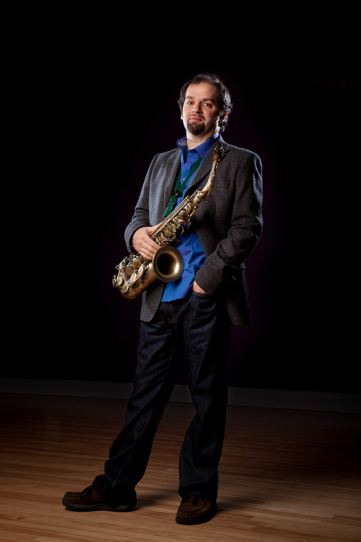 Jesse in the charcoal suit and blue shirt holding his saxophone in a classic pose. Dark shadowed background and wooden flooring.