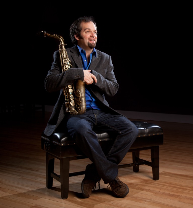 Jesse in the charcoal suit and blue shirt holding his saxophone sat on a piano stool. Dark shadowed background and wooden flooring.