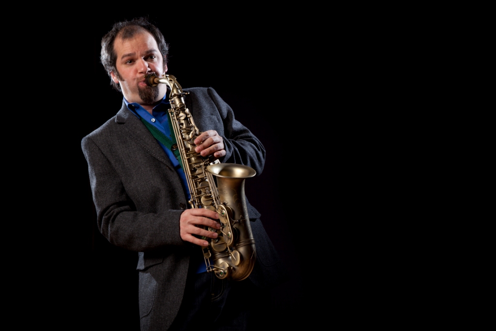 Jesse in charcoal suit and blue shirt stood playing the saxophone against a black background. 