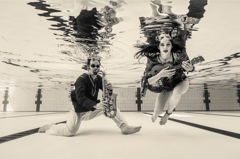 Jesse Bannister in red and white playing a glass saxophone with Maya Almeida in a multicoloured indian stitched top playing a blue ukelele. 