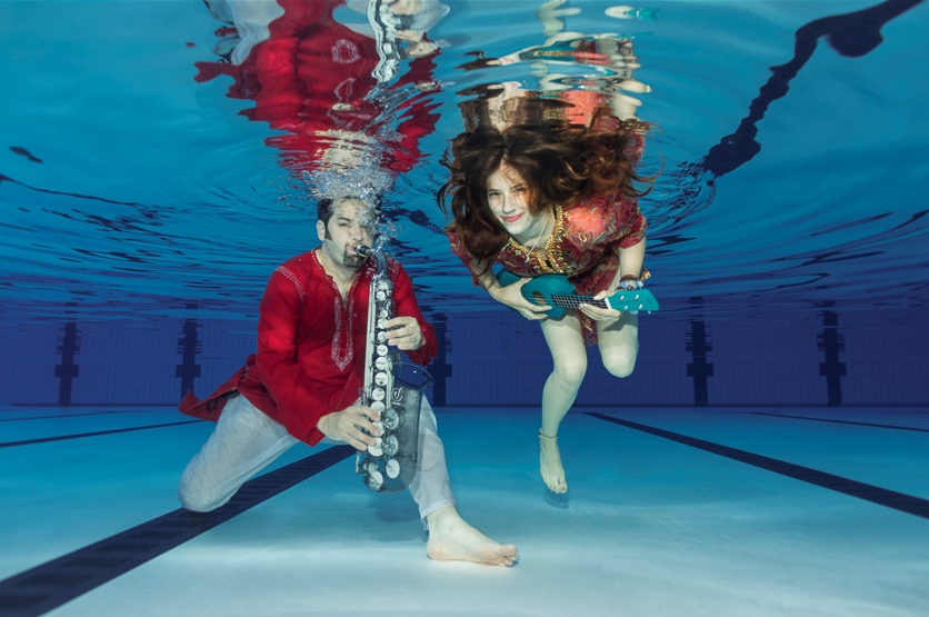 Jesse Bannister in red and white playing a glass saxophone with Maya Almeida in a multicoloured indian stitched top playing a blue ukelele.