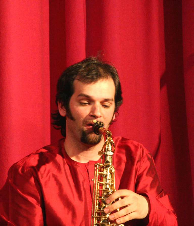Jesse Bannister in red playing the saxophone in front of a red curtain. 