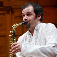 Jesse in a white shirt playing the saxophone. Internal wooden pannelling in the background.
