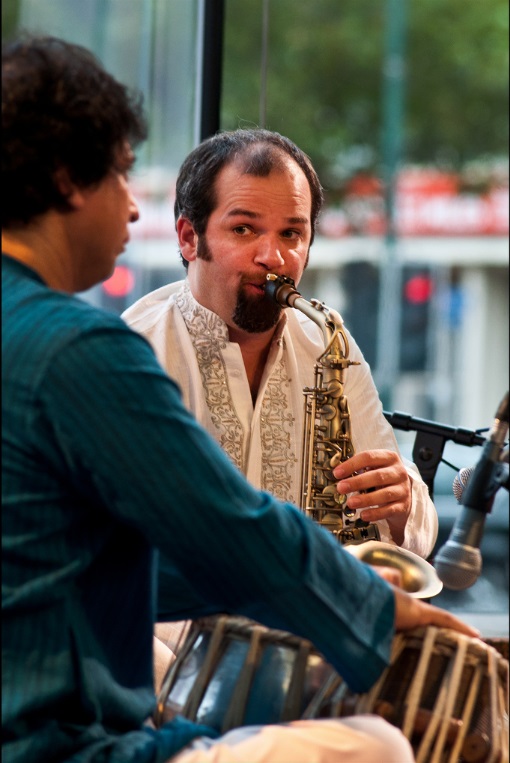 Jesse in a white punjabi suit, sat playing the saxophone with Sarvar Sabri in turquoise in the foreground.