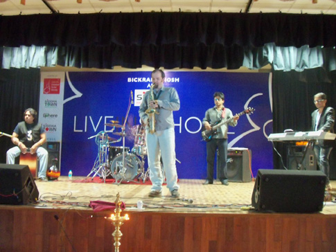 Jesse standing in a grey shirt playing the saxophone with the band and wall banner in the background.
