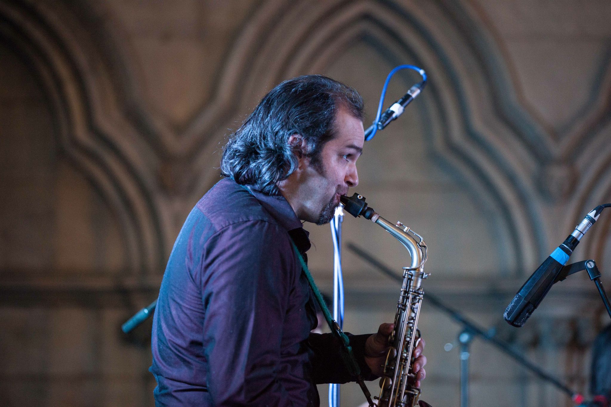Jesse in a purple shirt playing the saxophone in profil. Grey stone gothic arches of York Minister in the background/ 