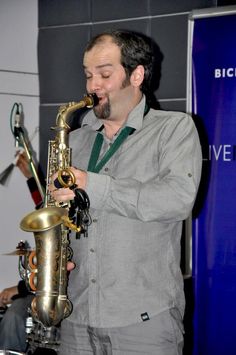 Jesse standing in a grey shirt playing the saxophone with event display boards in the background.